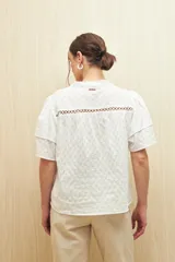 Camisa blanca de algodón liviano con cuello mao y cierre frontal de botones. Presenta un diseño de rayas verticales con textura y detalles de pasamanería calada en el frente y hombros, aportando un estilo romántico y artesanal.