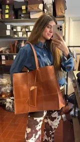 Bolso tote de cuero natural color camel, diseñado con piezas unidas mediante costuras visibles y terminaciones artesanales. Cuenta con doble asa de hombro y un pequeño estuche colgante decorativo.