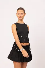 Musculosa negra de cuello redondo y calce entallado, decorada con alfileres de gancho plateados, cuentas negras y dijes metálicos dispuestos de forma irregular en el frente.