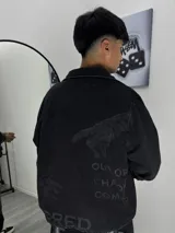 Campera de denim negro con diseño estampado en la espalda que incluye una mano y la frase 'OUT OF CHAOS COMES'. Presenta cuello camisero, cierre frontal y corte holgado.