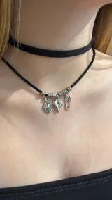 Gargantilla tipo choker de doble tira negra de gamuza, con dijes colgantes plateados en forma de brújula, corazón y alfiler de gancho, decorada con arandelas metálicas.