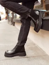 Botas cortas negras con plataforma dentada, confeccionadas en una combinación de cuero con textura de reptil y paneles de gamuza. Presentan un cierre frontal metálico y puntera cuadrada.
