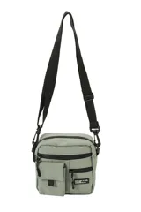 Morral de diseño compacto en color verde grisáceo, con correa ajustable y extraíble. Presenta un compartimento principal con cierre, un bolsillo frontal con solapa y otro bolsillo frontal con cierre, decorado con una etiqueta textil con texto.