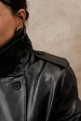 Trench largo de ecocuero negro, con cuello clásico, cierre cruzado con botones laterales y cinturón ajustable.