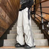 Pantalón deportivo unisex color beige, de corte ancho, con bolsillos laterales tipo cargo, cintura elástica ajustable con cordón, cierre lateral completo y detalle de tres rayas verticales negras en el lateral.