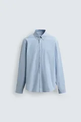 Camisa de hombre de manga larga, corte relajado (relaxed fit), color celeste con patrón de cuadros pequeños en azul y rojo. Presenta cuello abotonado (button-down), bolsillo de parche en el pecho y cierre frontal de botones.
