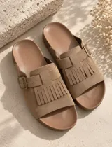 Sandalias de gamuza color marrón, con diseño de una sola tira ancha decorada con flecos y una hebilla metálica lateral ajustable. Cuenta con plantilla anatómica.