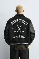 Cazadora estilo varsity con cuerpo de tejido con lana y mangas de efecto piel. Presenta cuello solapa, cierre frontal con botones a presión, bolsillos laterales y puños con elástico a rayas. Incluye parches y bordados decorativos en el frente.