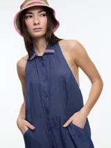 Vestido largo de lino en color azul, con cuello camisero, cierre frontal de botones y espalda descubierta con diseño estilo halter. Presenta un bordado decorativo en el cuello y bolsillos laterales.