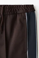 Pantalón deportivo marrón de punto con leve brillo, corte relajado y piernas rectas con pliegues cosidos. Cuenta con franjas laterales oscuras y ribetes blancos contrastantes, cintura elástica y bolsillos laterales.