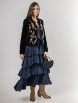 Chaqueta de terciopelo negro con bordados florales y ornamentales en tonos dorados, rosados y azules. Presenta un corte recto, cuello redondo y cierre frontal abierto con detalles de costuras decorativas en los bordes.