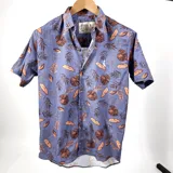 Camisa de manga corta con estampado de hojas en tonos marrones sobre fondo celeste. Tiene cuello italiano, cierre con botones y un bolsillo en el pecho con logo bordado.