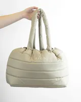 Bolso tipo tote color beige con diseño acolchado y asas tubulares. Presenta el logo de la marca en el centro.