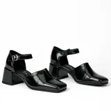 Zapato de cuero vacuno color gris topo, con talón descubierto y pulsera con hebilla. Tiene taco de madera forrado en cuero y suela de goma. La horma es de punta cuadrada, ancha y cómoda.