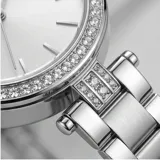 Reloj analógico de mujer con caja redonda de metal plateado y bisel decorado con cristales. Presenta una esfera minimalista con índices simples y una malla de eslabones de acero inoxidable con cierre desplegable.