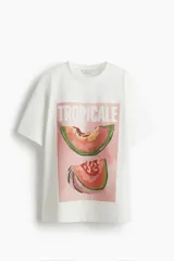 Remera oversized en punto de algodón, de color blanco, con cuello redondo, hombros caídos, mangas al codo y estampa frontal con la inscripción "TROPICALE" y gráficos de melón.