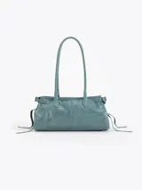 Cartera de hombro confeccionada en cuero vacuno color azul petróleo con acabado brillante. Presenta un diseño rectangular con herraje metálico tipo candado y dijes colgantes en el frente, además de correas laterales ajustables con tiras decorativas.