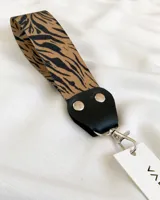 Llavero de tela con estampado animal print de cebra en tonos marrones y negros. Tiene un mosquetón metálico plateado y un aplique de cuero sintético negro con remaches plateados.
