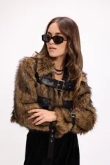 Campera corta de pelo sintético negro con detalles en cuero sintético, incluyendo bolsillos frontales con solapa, cinturón con hebilla metálica en el pecho y correas ajustables en los puños.