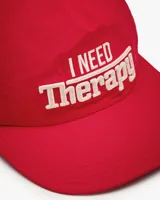 Gorra de béisbol de cinco paneles sin estructura color rojo, con visera plana y bordado frontal en tono crudo con la frase 'I NEED Therapy'. Presenta detalles bordados adicionales y correa ajustable de nailon en la parte trasera.