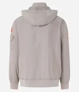 Campera rompevientos color beige con capucha, cierre frontal y bolsillos laterales. Presenta estampado de la NASA en el pecho, parche circular en la manga derecha y bandera de Estados Unidos en la manga izquierda.