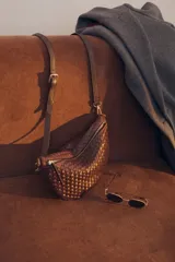 Bandolera de gamuza color marrón con diseño de media luna, decorada con tachas metálicas doradas distribuidas en el frente. Cuenta con cierre superior metálico y correa larga ajustable para llevar cruzada.