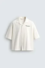 Camisa de corte relajado confeccionada en algodón, con cuello bowling y manga corta. Presenta cierre frontal con botones y detalle de texto estampado en el pecho.