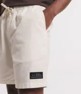 Bermuda masculina de sarga color blanco, con cintura elástica y lazo de ajuste. Presenta bolsillos laterales y una etiqueta decorativa aplicada en la parte inferior de la pierna.