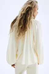 Blusa de manga globo larga color crema, con cuello redondo fruncido y botones ocultos en la parte delantera. Presenta un bajo redondeado.