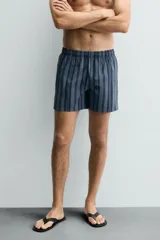 Bañador de hombre de largo medio, color azul claro con rayas verticales en azul oscuro. Posee cintura elástica ajustable con cordón, bolsillos laterales y un bolsillo trasero de plastrón.