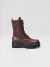 Botas de cuero color marrón de caña alta, con plataforma gruesa de goma y cierre metálico frontal con tirador. Presentan costuras a contratono y detalle de etiqueta de la marca en la lengüeta.