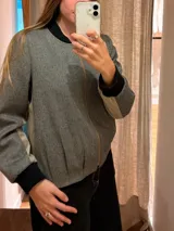 Chaqueta tipo bomber de lana con estampado de espiga en tonos grises. Presenta cuello, puños y ruedo en tejido elástico negro, cierre frontal dorado y bordado sutil en el pecho.