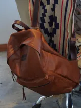 Mochila de cuero negro, con forro interior de lino y herrajes de bronce. Presenta un bolsillo frontal con cierre y un parche de cuero marrón con logo grabado.