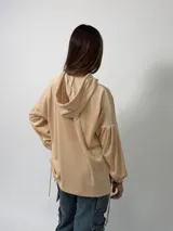 Canguro de plush color beige con capucha, bolsillo frontal tipo canguro, mangas globo con puños fruncidos y tajos laterales con tiras ajustables.