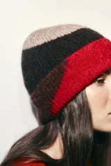 Gorro tipo beanie de punto con diseño de bloques de color en tonos rojo, negro y beige. Presenta un acabado con vuelta y textura suave.
