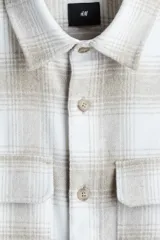 Sobrecamisa de franela con cuello inglés, tapeta lisa, dos bolsillos superiores con solapa y mangas largas con puños abotonados. Presenta un patrón de cuadros en tonos blanco y beige/tostado.