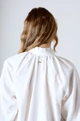 Blusa blanca de algodón con cuello alto con volantes y botonadura frontal parcial. De manga larga con puños abullonados.
