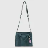 Bolso tipo tote mini de la marca Bubba, color verde oscuro, con doble asa de mano y correa larga ajustable y extraíble. Presenta el logo de la marca en el frente y un dije colgante con la letra B en color rosado.