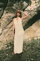 Vestido midi de color beige con escote en pico y tirantes anchos. Presenta un diseño con tejido plisado en la parte superior y aplicaciones de encaje a tono en el escote y la cintura. Cuenta con una tira ajustable con lazada en la espalda y forro interior.