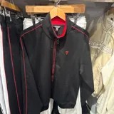Campera deportiva Guess de color negro, con cierre frontal completo, cuello alto y detalles de líneas rojas en los laterales y el cuello. Presenta el logo triangular de la marca bordado en el pecho.