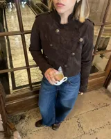 Campera de estilo militar en color marrón oscuro, confeccionada en gamuza. Presenta un diseño corto, cuello alto y cierre frontal cruzado con botones metálicos decorativos. Incluye detalles de charreteras en los hombros y botones en los puños.