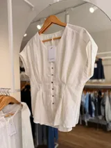 Blusa blanca de lino con escote en V, manga corta caída y botonadura frontal con botones oscuros. Presenta pinzas en la cintura que definen la silueta.