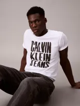 Remera blanca de algodón con cuello redondo y manga corta, presentando un estampado gráfico apilado en negro con el texto "CALVIN KLEIN JEANS".