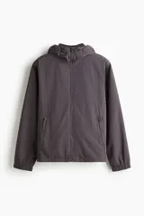 Campera rompevientos de nylon repelente al agua, color gris oscuro/marrón topo, con forro de malla. Presenta cierre frontal con protección para el mentón, capucha ajustable con cordón, bolsillos laterales y bolsillo interior con cierre, y elástico revestido en puños y bajo.