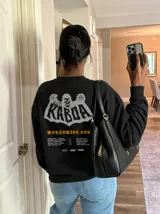 Buzo gris oscuro con efecto acid wash, corte regular fit y cuello redondo. Presenta una estampa grande en la espalda con diseño de calavera, el texto "Kaboa" y detalles de fechas de tour en tonos claros.
