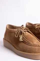 Zapato tipo mocasín acordonado de gamuza color marrón oscuro, con suela de goma color caramelo y detalle de dije metálico dorado en forma de bota colgando de los cordones.