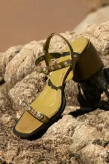 Sandalia de cuero color verde oliva con taco medio y punta cuadrada. Presenta tiras adornadas con tachas esféricas plateadas, una tira delantera y una tira de tobillo ajustable con hebilla.