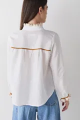 Camisa de manga larga en color blanco, confeccionada en tejido de lino, con detalles de volados en crochet en el cuello, la botonadura frontal y los puños. Presenta detalles de costuras en color ocre en los hombros y puños, con cierre frontal de botones.