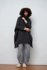 Poncho de tejido artesanal en color negro, con cuello alto tipo polera, mangas largas y terminación con flecos en el ruedo. Presenta una etiqueta de cuero decorativa en la parte inferior.
