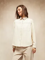 Camisa de manga larga color crudo, con cuello clásico, botones frontales y bolsillo de parche en el pecho. Presenta costuras decorativas en contraste.